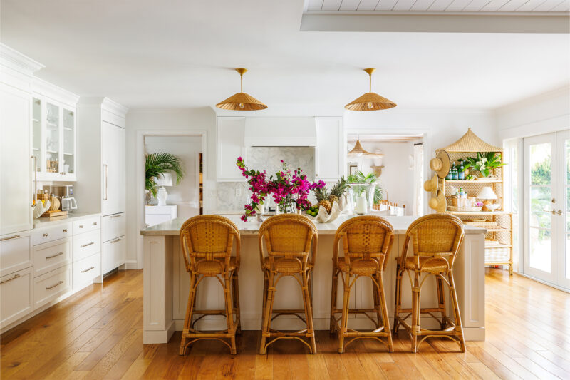 A fresh white kitchen with a light & airy feel. Bamboo barstools are placed in front of a large kitchen island, with rattan light fixtures floating above. Bougainvillea cuttings & pineapple are on the countertop.